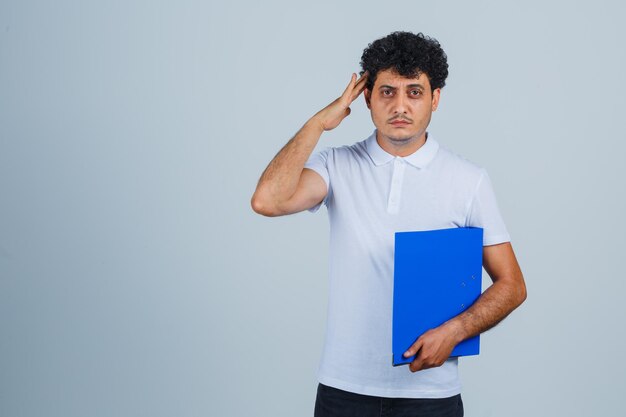 Young man holding file folder, showing salute gesture in white t-shirt and jeans and looking serious. front view.