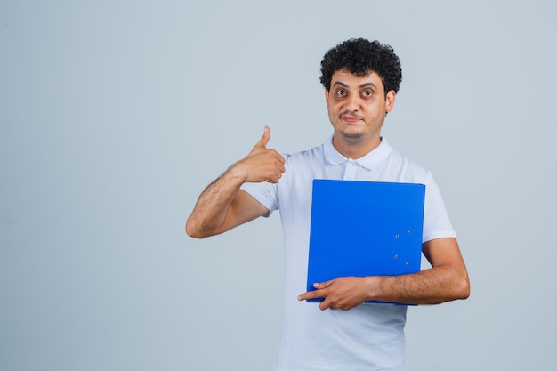 Молодой человек держит папку с файлами и показывает большой палец вверх в белой футболке и джинсах и выглядит счастливым, вид спереди.