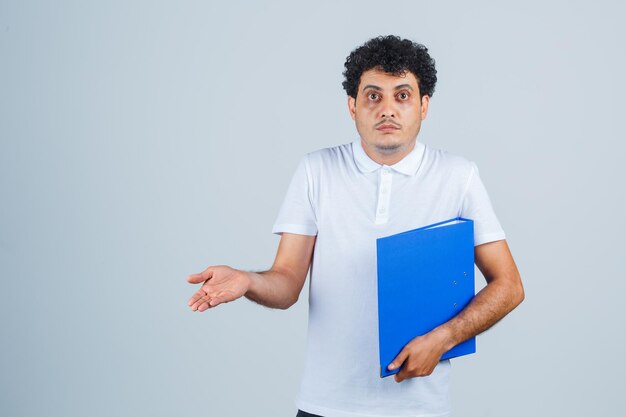 Young man holding file folder and stretching hand toward camera in white t-shirt and jeans and looking perplexed. front view.