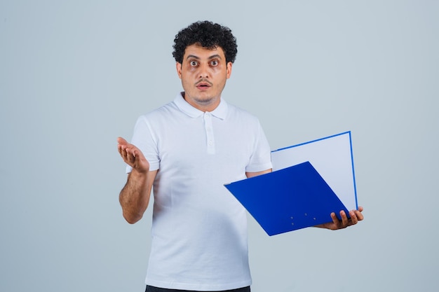 Young man holding file folder and stretching hand toward camera in white t-shirt and jeans and looking surprised , front view.