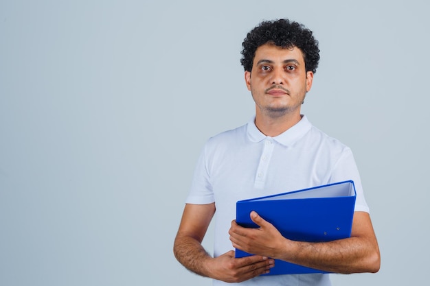 Young man holding file folder in white t-shirt and jeans and looking serious , front view.