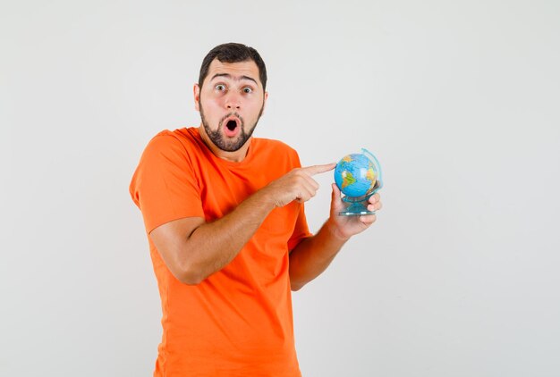 Young man holding finger on globe in orange t-shirt and looking surprised , front view.