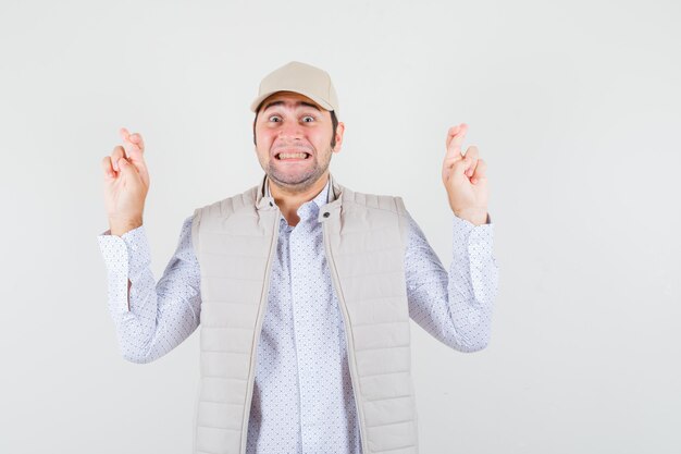 Young man holding fingers crossed in beige jacket and cap and looking happy , front view.