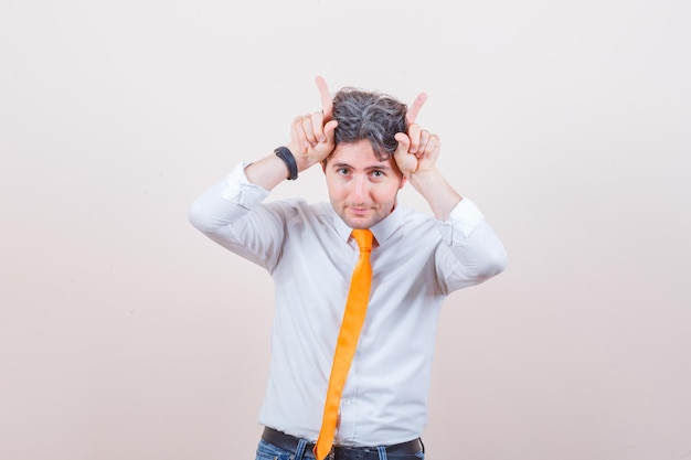 Young man holding fingers over head as bull horns in shirt, jeans and looking funny