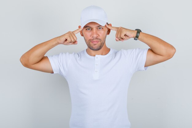 Young man holding fingers on his temples in white t-shirt, cap, front view.