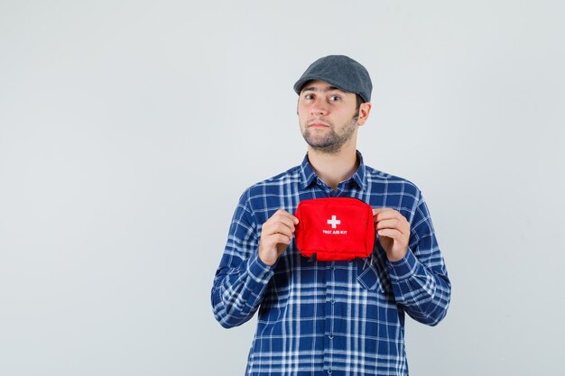 Young man holding first aid kit in shirt, cap and looking confident , front view.