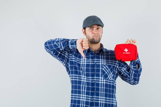 Young man holding first aid kit, showing thumb down in shirt, cap front view.