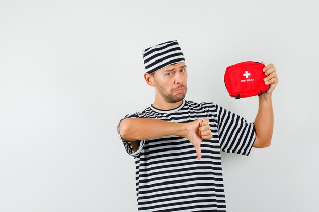 Young man holding first aid kit showing thumb down in striped t-shirt hat and looking disappointed 