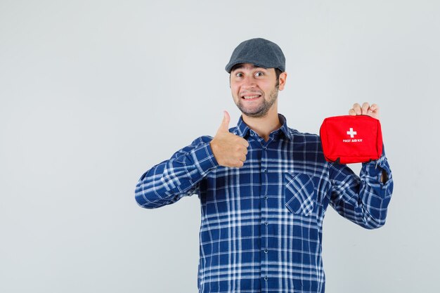 Young man holding first aid kit, showing thumb up in shirt, cap and looking cheery , front view.