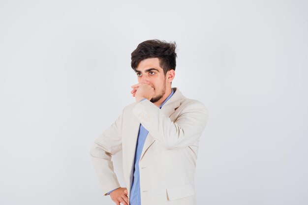 Молодой человек держит кулак во рту, держа другую руку на талии в синей рубашке и белом пиджаке и выглядит задумчиво