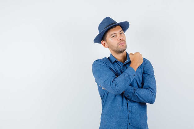 Giovane che tiene il pugno alzato in camicia blu, cappello e sembra sicuro. vista frontale.