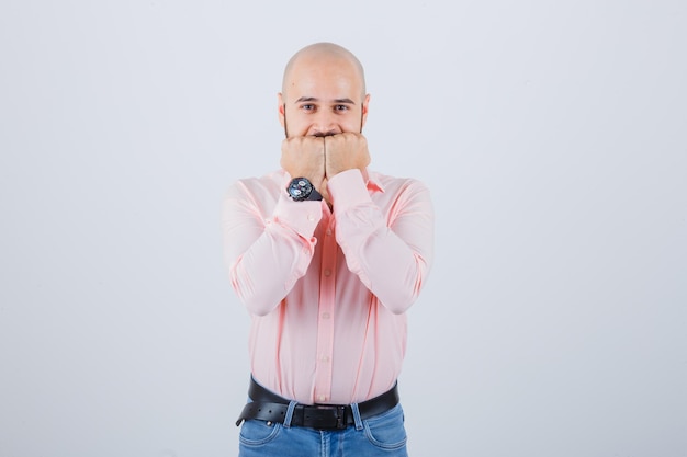 Young man holding fists on mouth in pink shirt,jeans and looking happy , front view.