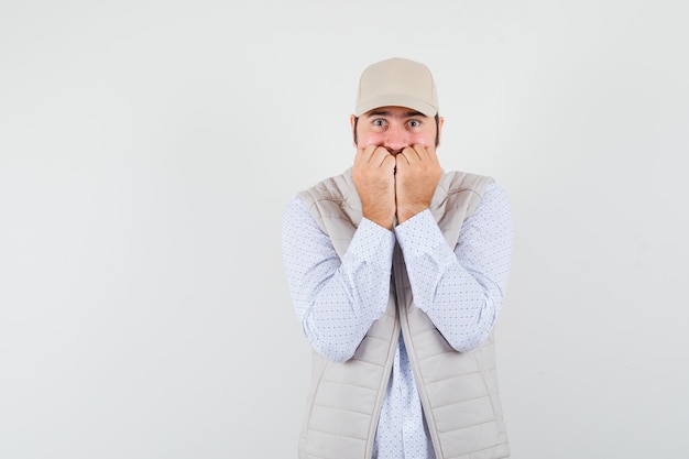 Free photo young man holding fists on mouth in shirt,sleeveless jacket,cap and looking troubled , front view. space for text