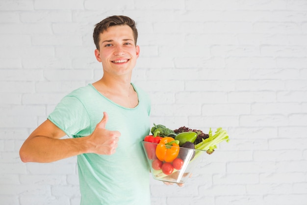 Молодой человек, держащий свежие овощи в стеклянной чаше, показывая пальцем вверх знак
