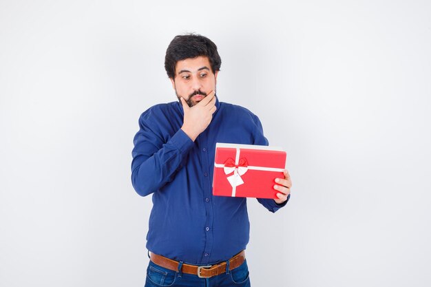 Young man holding gift box and putting hand on chin in blue shirt and jeans and looking pensive. front view.