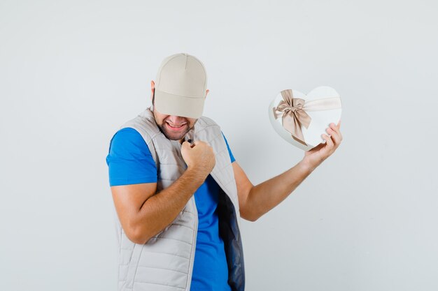 Young man holding gift box in t-shirt, jacket and looking blissful. front view.