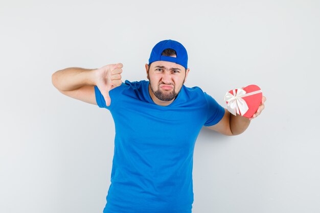 Молодой человек держит подарочную коробку большим пальцем вниз в синей футболке и кепке и выглядит задумчиво