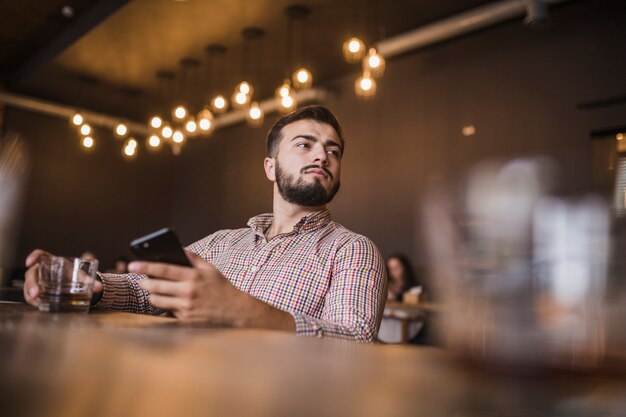 Young man holding glass of drink and mobile phone looking away