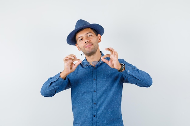 Young man holding glasses in blue shirt, hat and looking cheerful. front view.