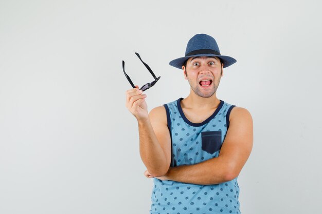 Young man holding glasses in blue singlet, hat and looking astonished. 