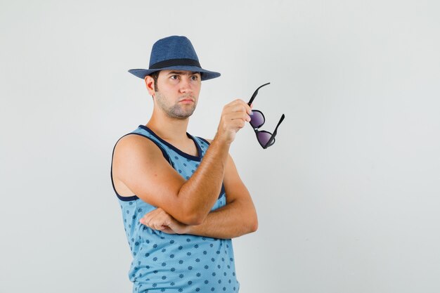 Молодой человек держит очки в синей майке, шляпе и выглядит задумчивым