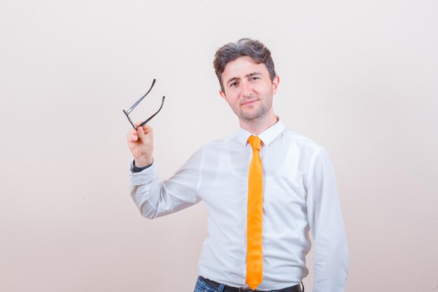 Young man holding glasses in shirt, jeans and looking jolly