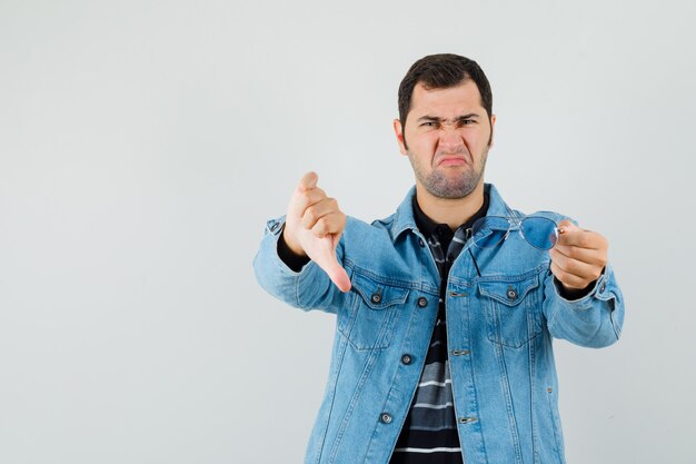 Young man holding glasses, showing thumb down in t-shirt, jacket and looking displeased. 