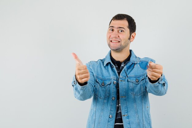 Young man holding glasses, showing thumb up in t-shirt, jacket and looking pleased