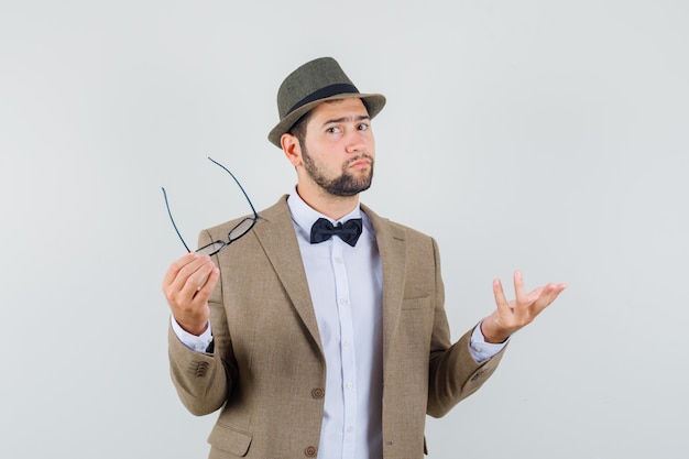 Free photo young man holding glasses in suit, hat and looking puzzled , front view.