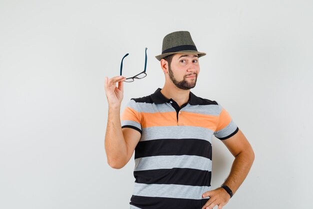 Young man holding glasses in t-shirt, hat and looking optimistic , front view.