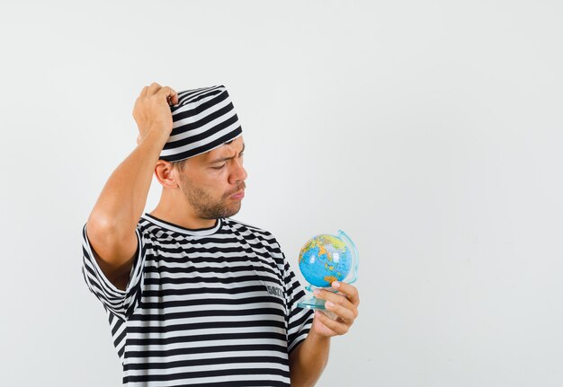 Young man holding globe while thinking in striped t-shirt hat and looking careful 