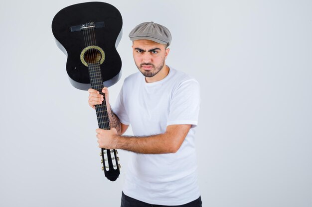 Молодой человек держит гитару в белой футболке, черных штанах, серой кепке и выглядит разъяренным