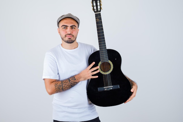 Young man holding guitar in white t-shirt, black pants, grey cap and looking serious