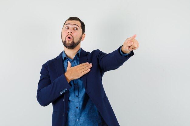 Young man holding hand on chest, pointing away in shirt, jacket front view.