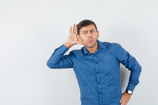 Young man holding hand behind ear in blue shirt and looking snoopy , front view.