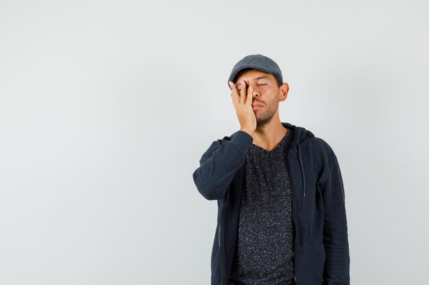 Young man holding hand on face in t-shirt, jacket, cap and looking sleepy. front view.