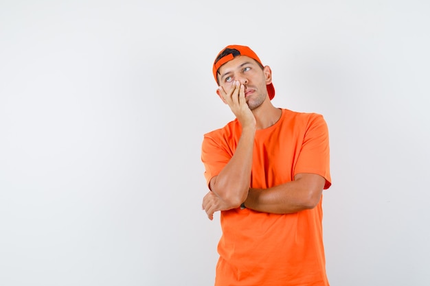 Young man holding hand on face while thinking in orange t-shirt and cap and looking exhausted