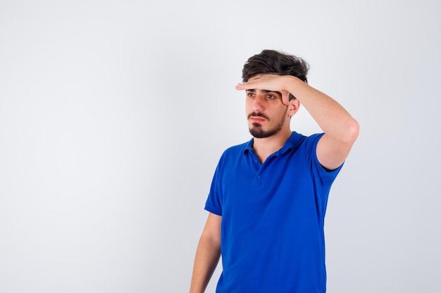 Young man holding hand on forehead, looking away in blue t-shirt and looking serious