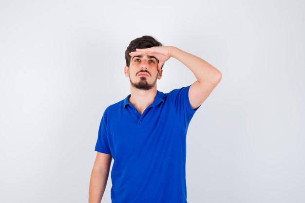 Young man holding hand on forehead, looking away in blue t-shirt and looking serious