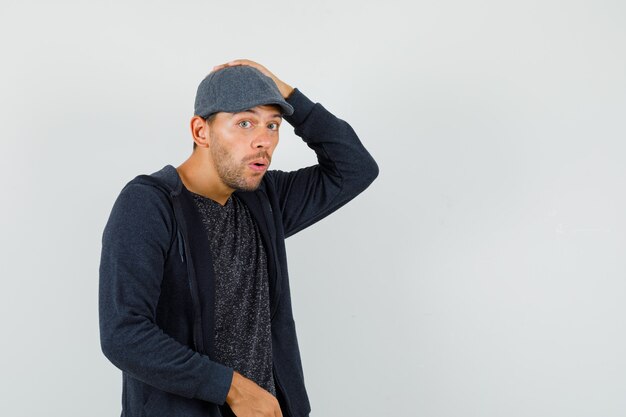 Young man holding hand on head in t-shirt, jacket, cap and looking surprised. front view.