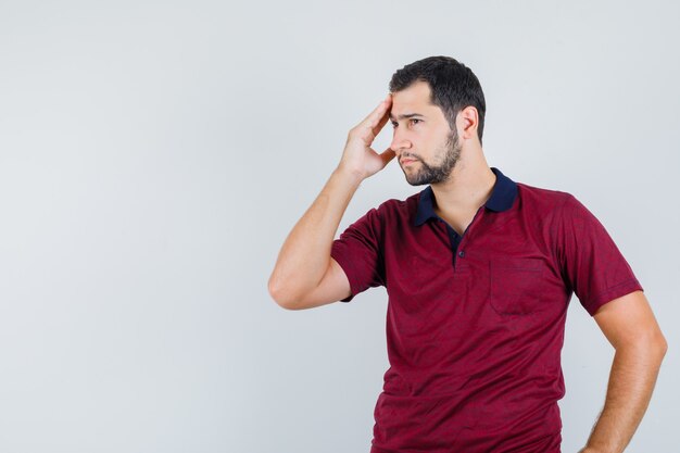 Young man holding hand on his forehead in red t-shirt and looking thoughtful. front view.