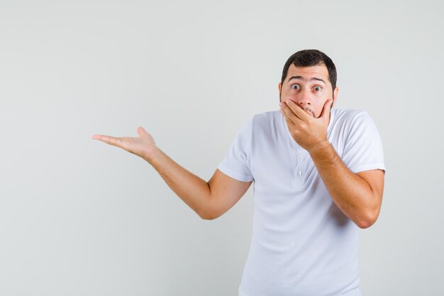 Young man holding hand on his mouth while pointing aside in t-shirt and looking amazed , front view.