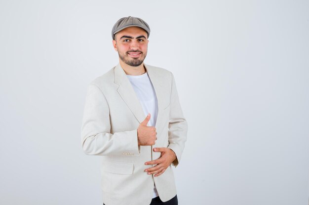 Young man holding hand on jacket while showing thumb up in white t-shirt, jacket and grey cap and looking happy