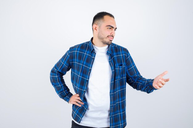 Young man holding hand on waist while stretching hand as holding something in checked shirt and white t-shirt and looking happy