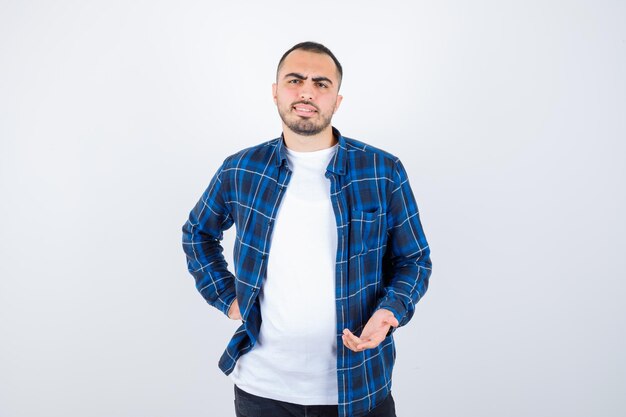 Young man holding hand on waist while stretching hand as holding something in checked shirt and white t-shirt and looking happy