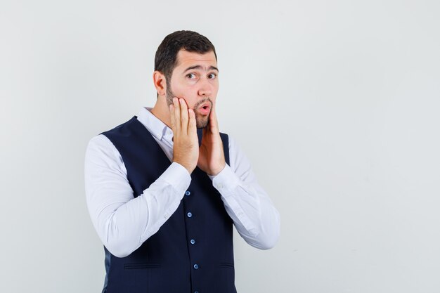 Young man holding hands on cheeks in shirt, vest and looking surprised