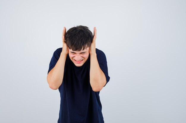 Young man holding hands on ears in black t-shirt and looking irritated. front view.