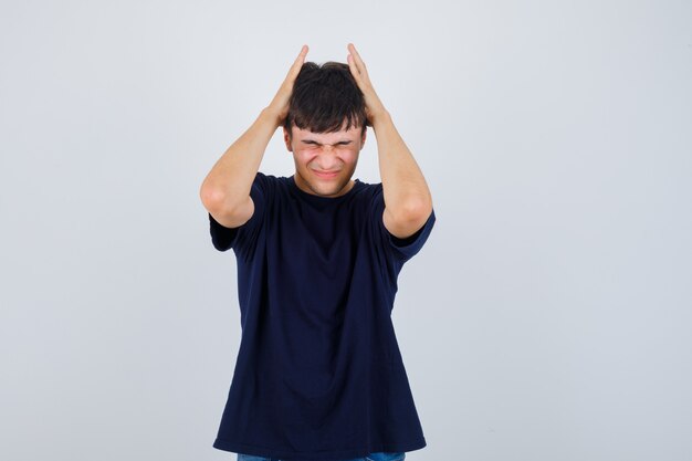 Young man holding hands on head in black t-shirt and looking irritated , front view.