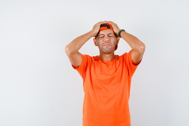 Молодой человек, держащий руки на голове в оранжевой футболке и кепке и выглядящий с сожалением
