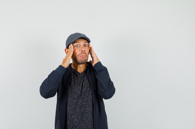 Young man holding hands to head in t-shirt, jacket, cap and looking confused , front view.